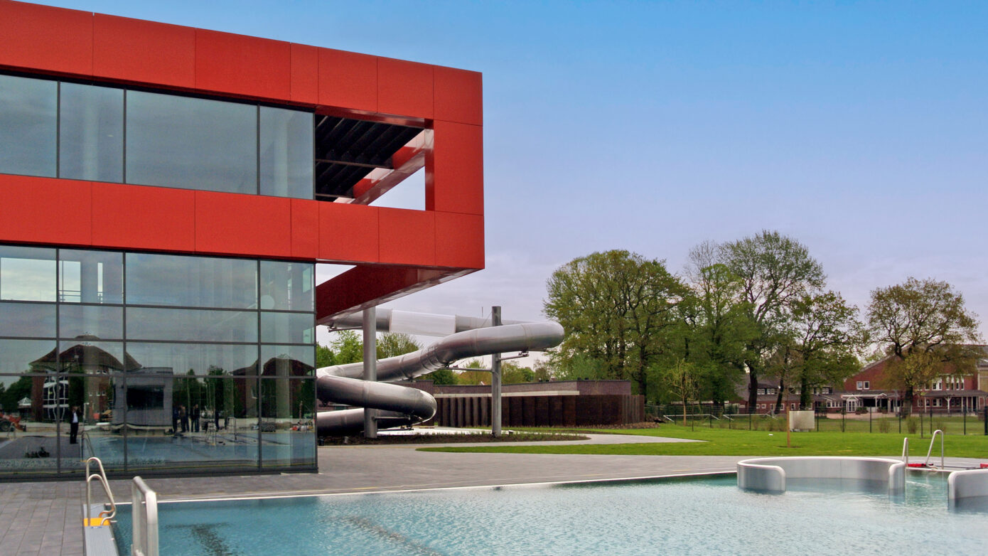 Moderne Schwimmhalle mit Rutsche und großzügigem Außenbereich in einer grünen Umgebung.