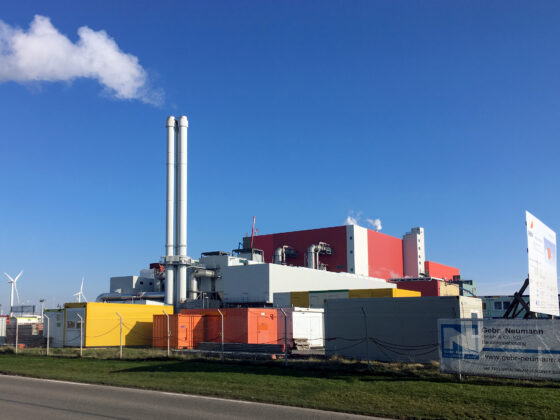Ersatzbrennstoff-Heizkraftwerk Delfzijl mit Schornsteinen, Windkraftanlagen und bunten Containern unter blauem Himmel.