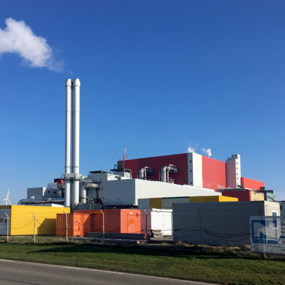 Ersatzbrennstoff-Heizkraftwerk Delfzijl mit Schornsteinen, Windkraftanlagen und bunten Containern unter blauem Himmel.