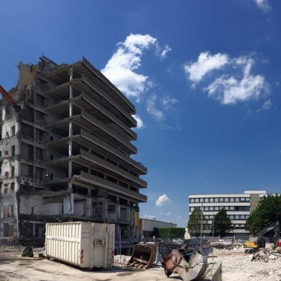 Abgerissenes Gebäude mit Bagger, Baustelle und blauen Himmel, Wolken im Hintergrund.