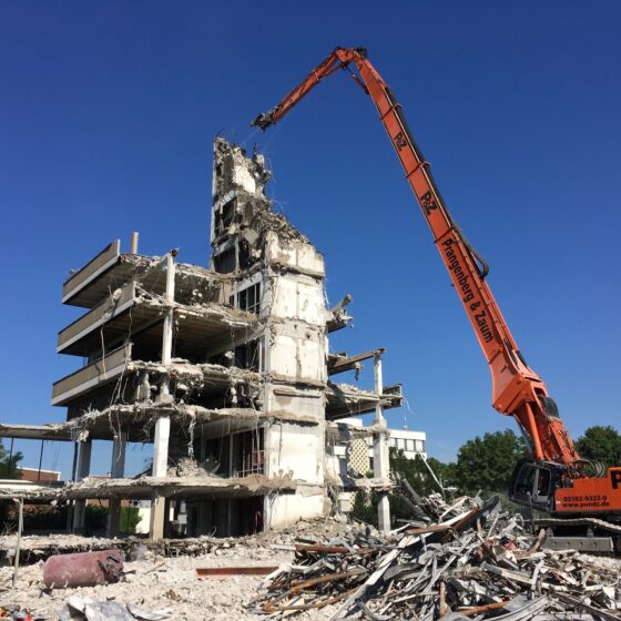 Bagger demoliert ein Gebäude unter klarem blauen Himmel, Baustelle im Hintergrund.
