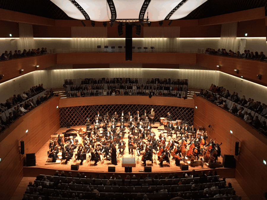 Orchesteraufführung in modernem Konzertsaal mit Publikum auf mehreren Ebenen.