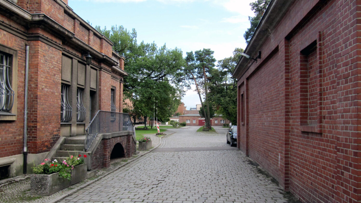 Pflasterstraße zwischen historischen Backsteingebäuden mit Bäumen im Hintergrund.