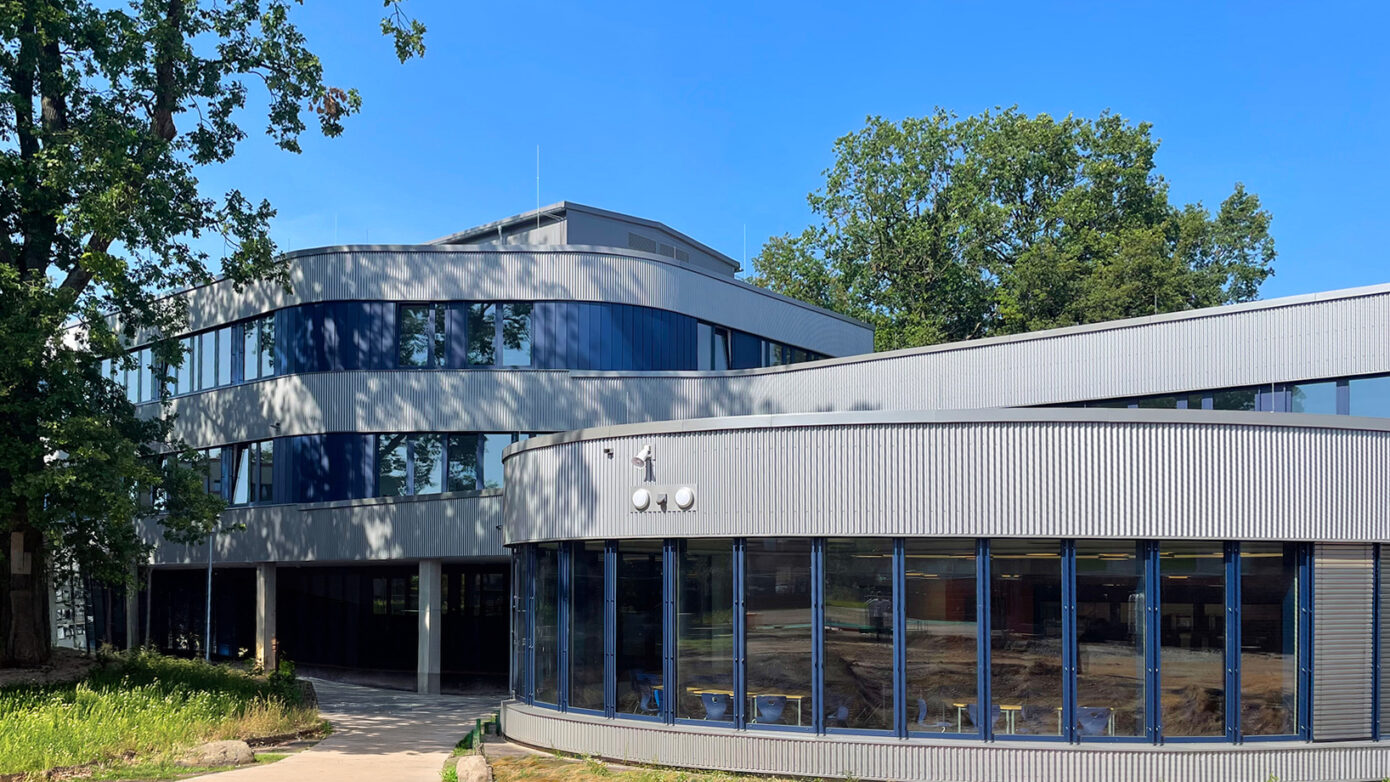 Moderne Architektur mit großen Fenstern und grünem Umfeld unter blauem Himmel.