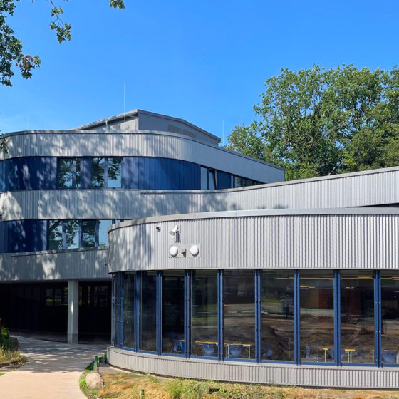 Moderne Architektur mit großen Fenstern und grünem Umfeld unter blauem Himmel.