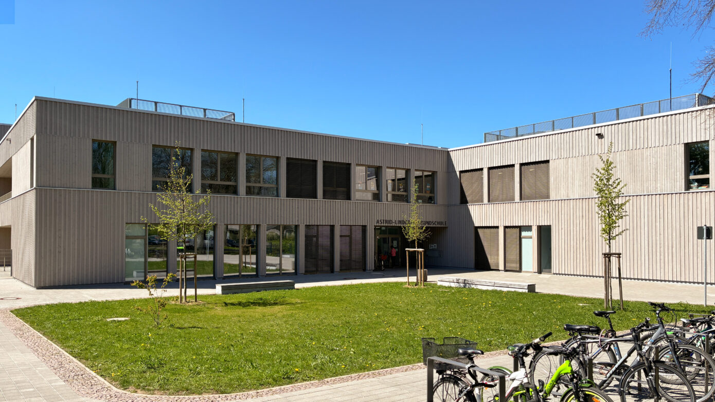 Schulgebäude der Astrid-Lindgren-Grundschule mit Holzfassade und Wiese und Fahrrädern im Vordergrund an sonnigem Tag.