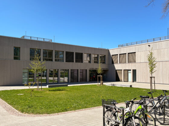 Schulgebäude der Astrid-Lindgren-Grundschule mit Holzfassade und Wiese und Fahrrädern im Vordergrund an sonnigem Tag.