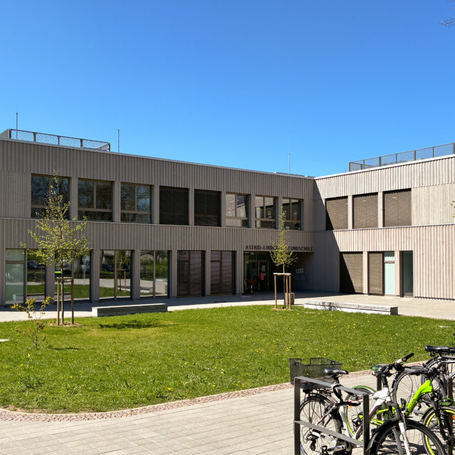 Schulgebäude der Astrid-Lindgren-Grundschule mit Holzfassade und Wiese und Fahrrädern im Vordergrund an sonnigem Tag.