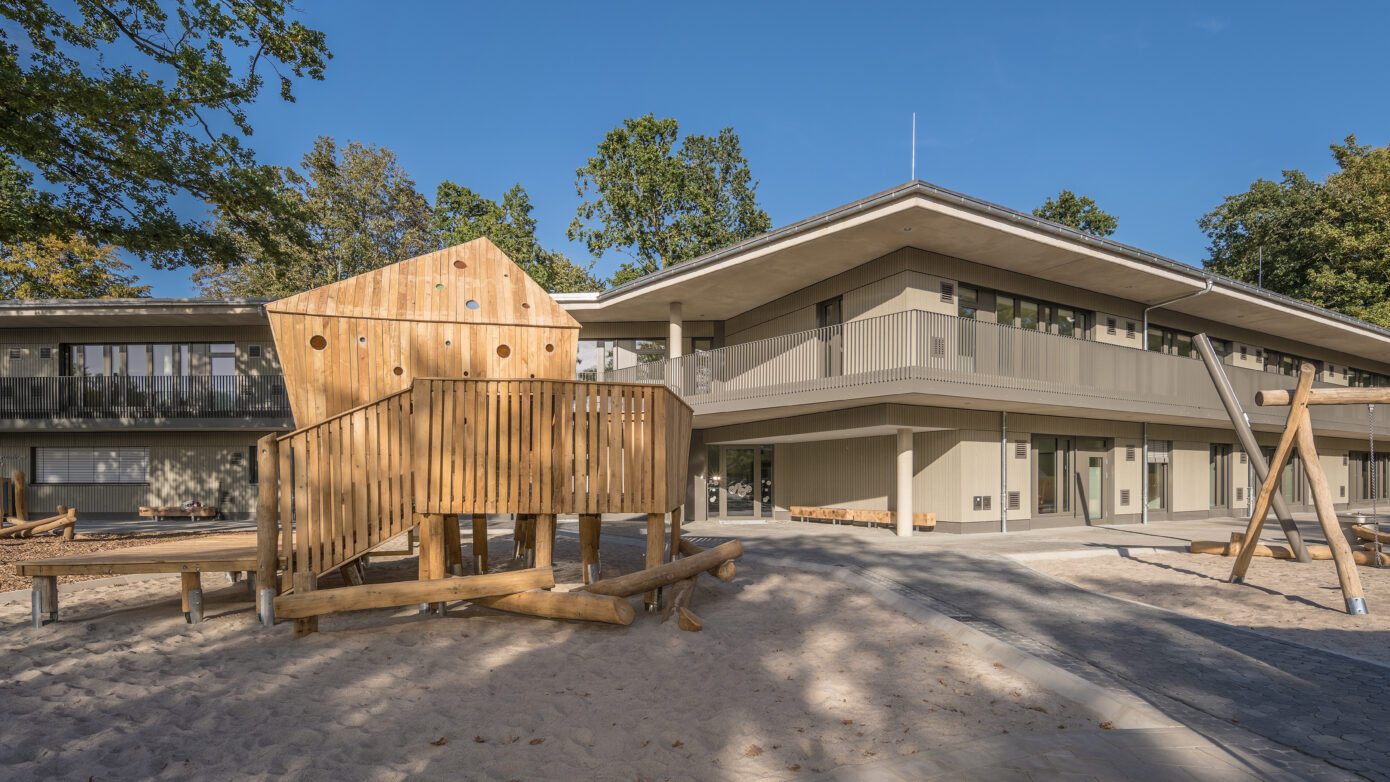 Moderne Kita mit Spielplatz und Holzspielgeräten in grüner Umgebung.