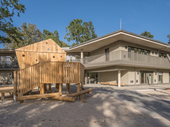 Moderne Kita mit Spielplatz und Holzspielgeräten in grüner Umgebung.