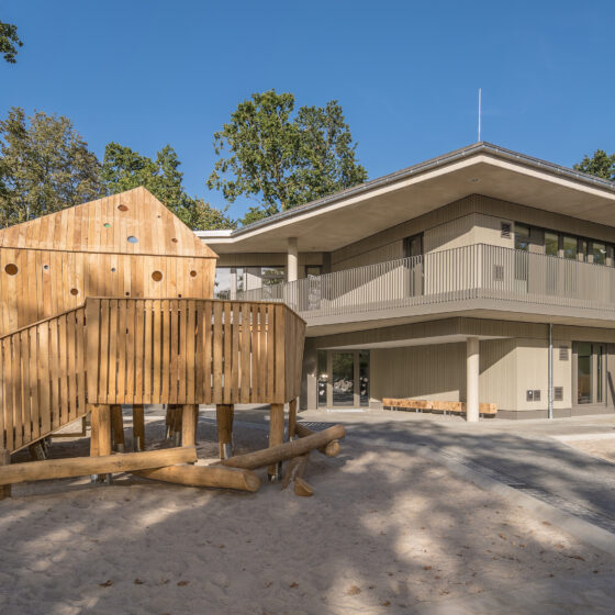 Moderne Kita mit Spielplatz und Holzspielgeräten in grüner Umgebung.