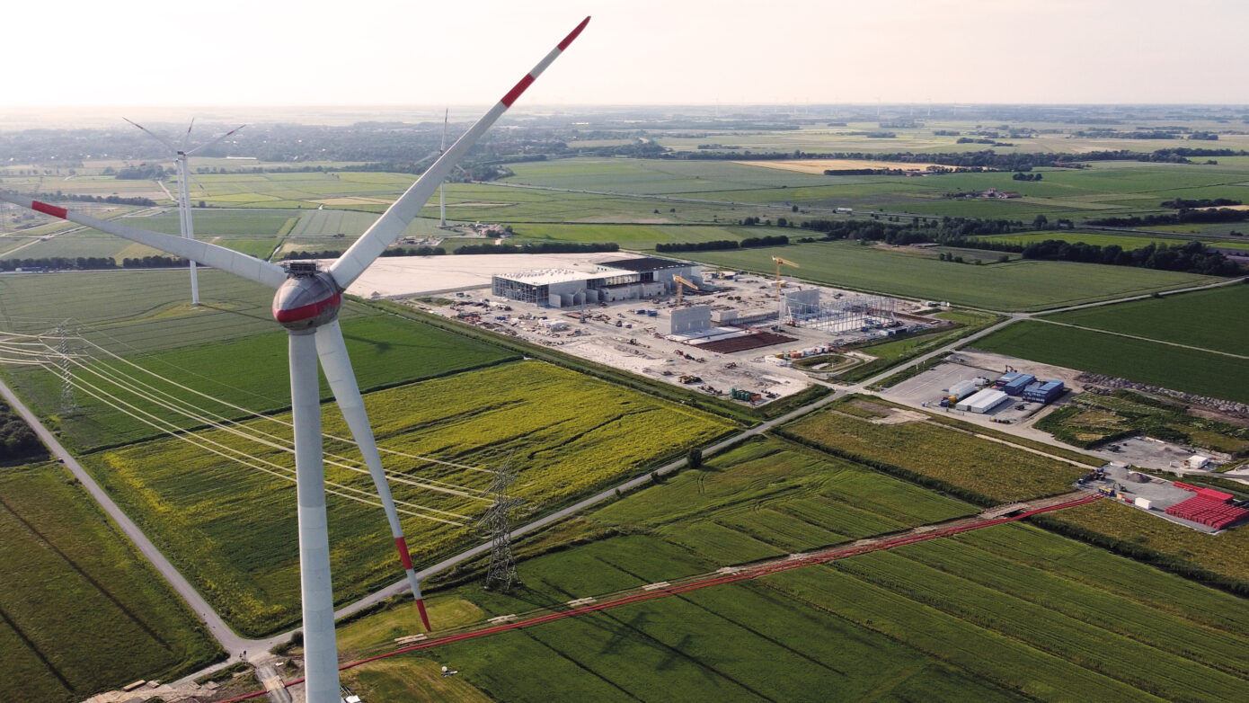 Windkraftanlage über grünen Feldern und einem Industriekomplex in ländlicher Umgebung.