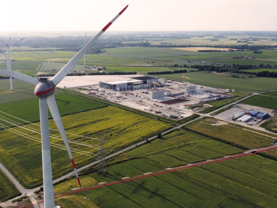 Windkraftanlage über grünen Feldern und einem Industriekomplex in ländlicher Umgebung.