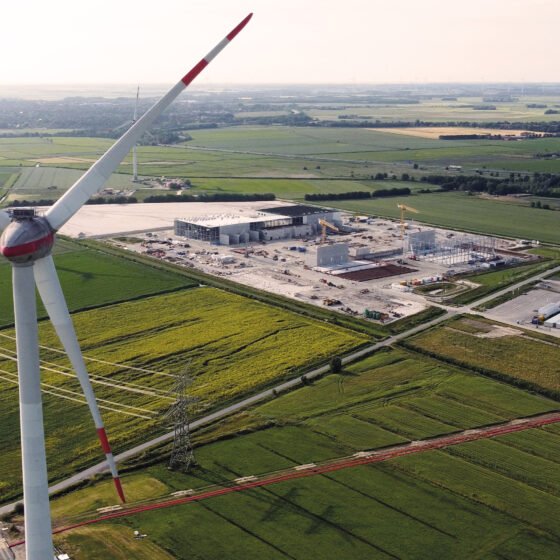 Windkraftanlage über grünen Feldern und einem Industriekomplex in ländlicher Umgebung.