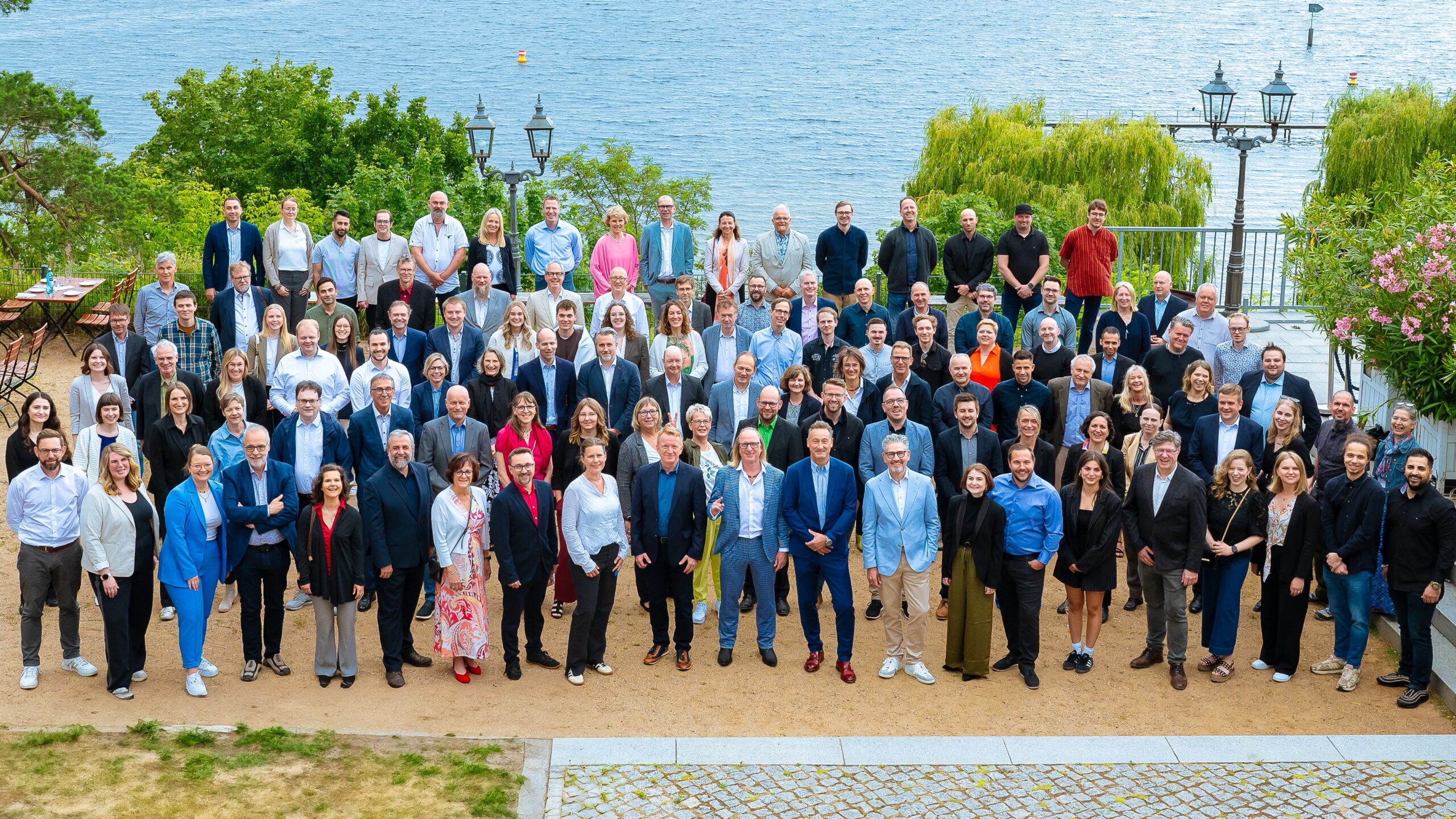 Gruppenfoto von über 100 Personen am Wasser, umgeben von Bäumen und Laternen.