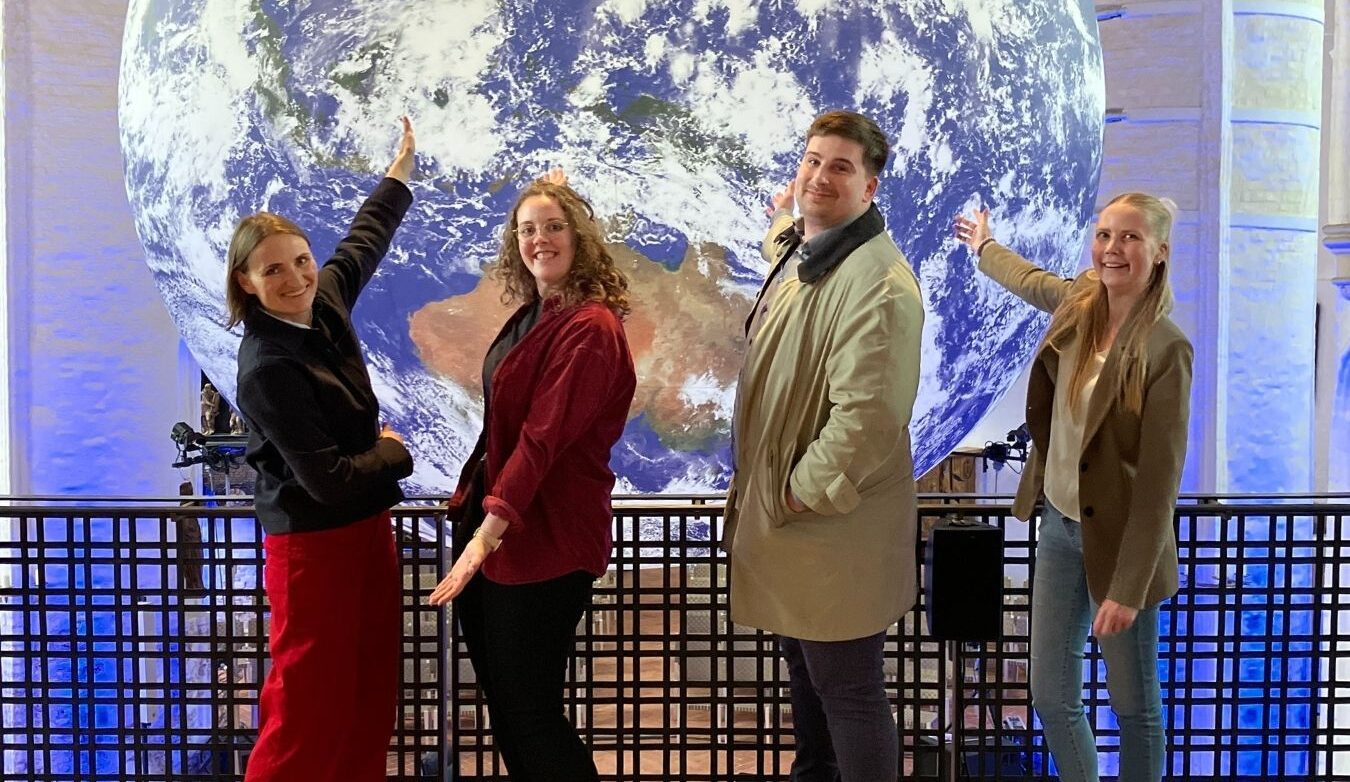 Gruppe von vier Personen posiert vor einer großen Erdkugel in einer modernen Ausstellung.