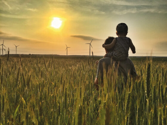 Vater mit Kind auf den Schultern im Weizenfeld bei Sonnenuntergang und Windrädern.