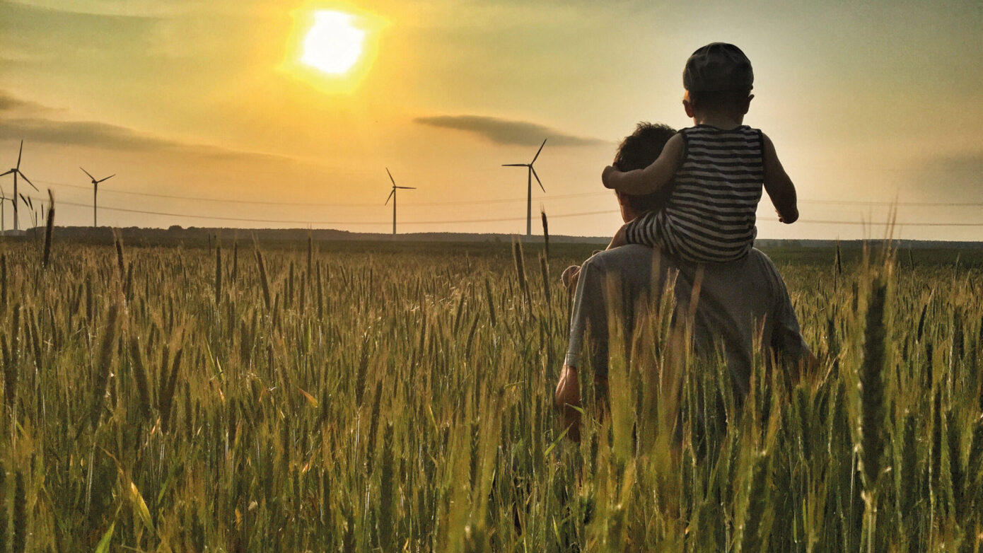 Vater mit Kind auf den Schultern im Weizenfeld bei Sonnenuntergang und Windrädern.