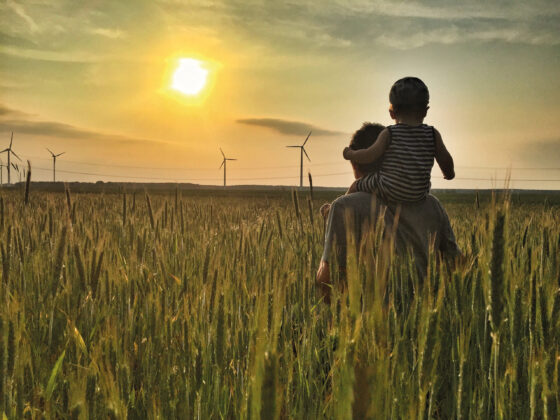Vater mit Kind auf den Schultern im Weizenfeld bei Sonnenuntergang und Windrädern.