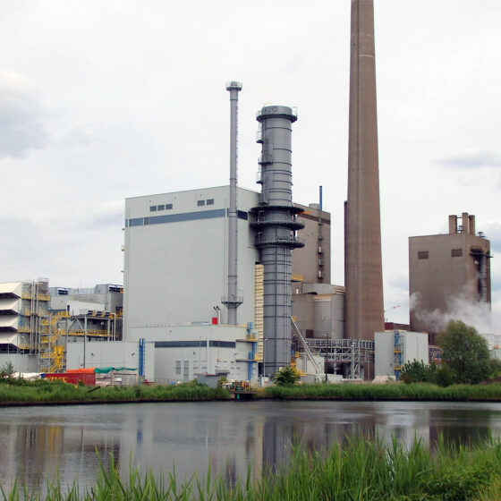 GUD-Kraftwerk mit Kühlturm und Produktionsgebäude am Wasser, umgeben von grüner Vegetation.