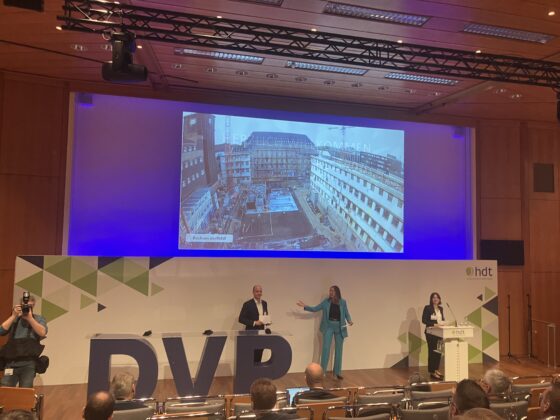 Konferenzszene mit Präsentation und Publikum, moderne Architektur im Hintergrund.