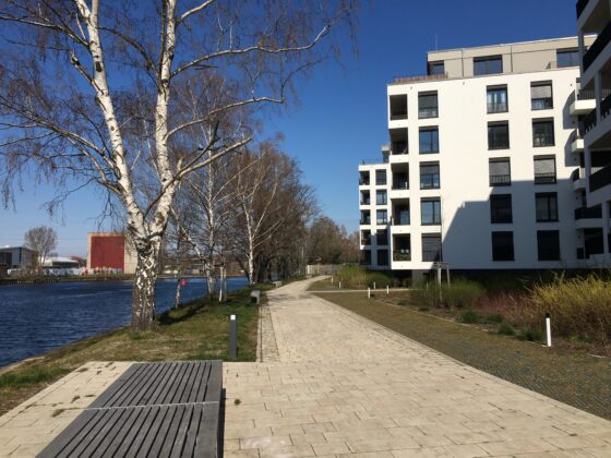 Moderne Wohnanlage am Wasser mit Gehweg und Bäumen unter blauem Himmel.