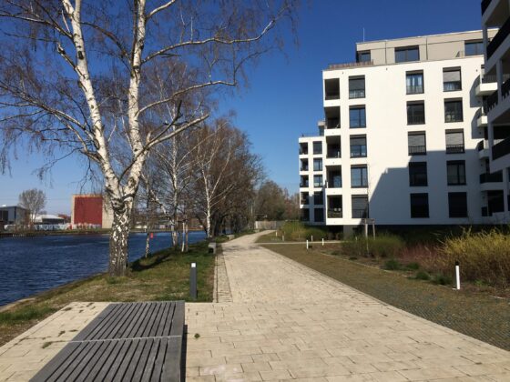 Moderne Wohnanlage am Wasser mit Gehweg und Bäumen unter blauem Himmel.