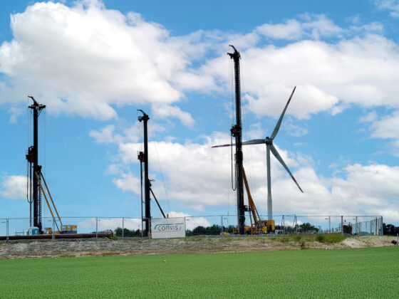 Bauarbeiten mit Kränen und Windkraftanlage vor einem blauen Himmel und grünen Feldern.