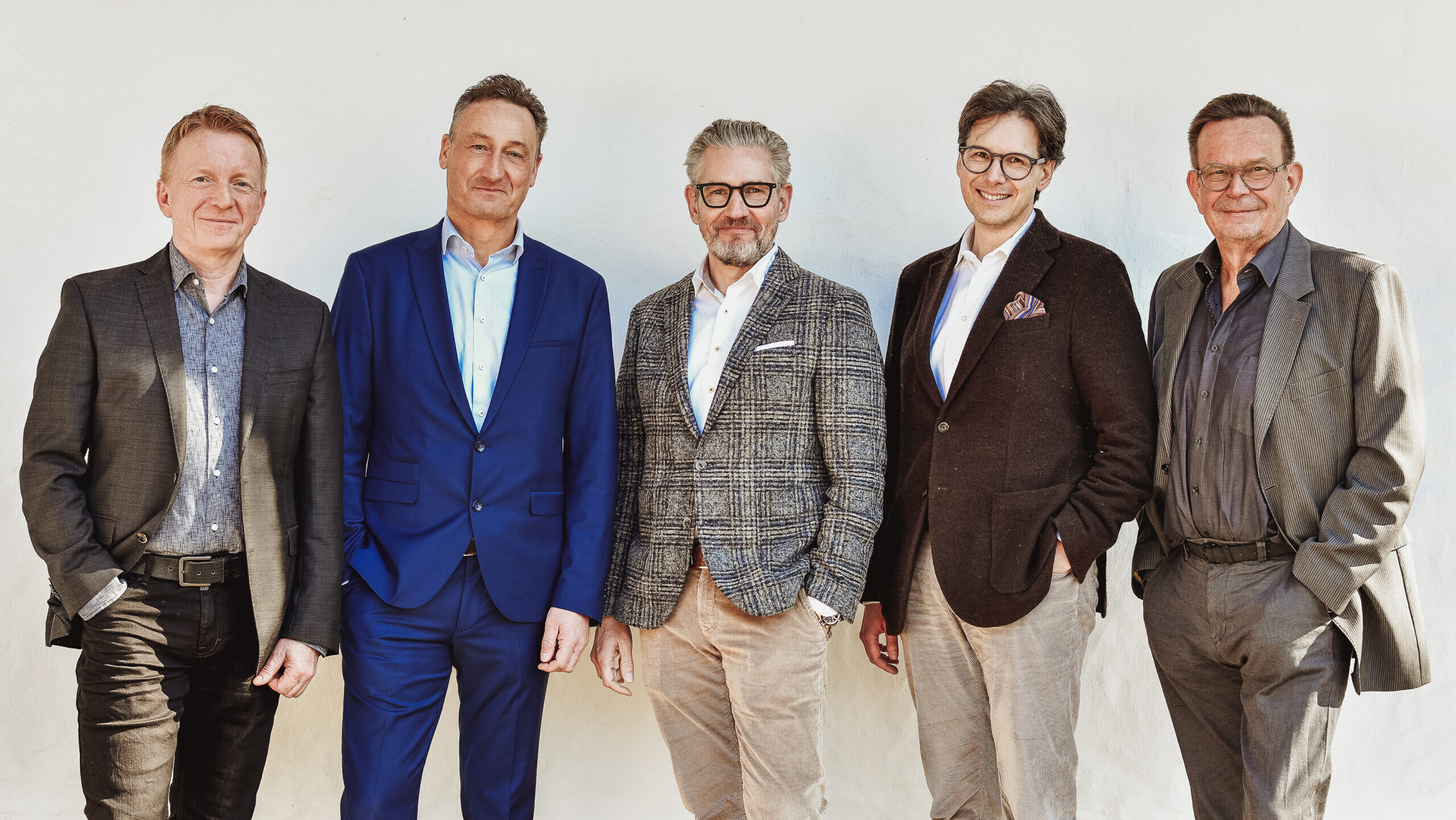 Fünf Männer in Anzügen stehen vor einer weißen Wand und lächeln freundlich.