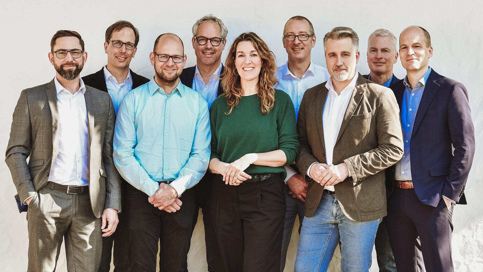 Gruppenfoto von neun Personen in formeller Kleidung vor einer weißen Wand.