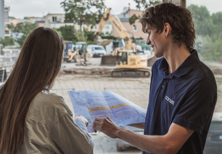 Baustellenbesprechung: Fachkräfte prüfen Baupläne vor einem Erdbaumaschinen-Hintergrund.