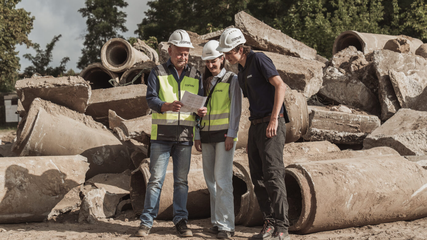 Bauarbeiter besprechen Pläne vor einer Baustelle mit Betonrohren im Hintergrund.