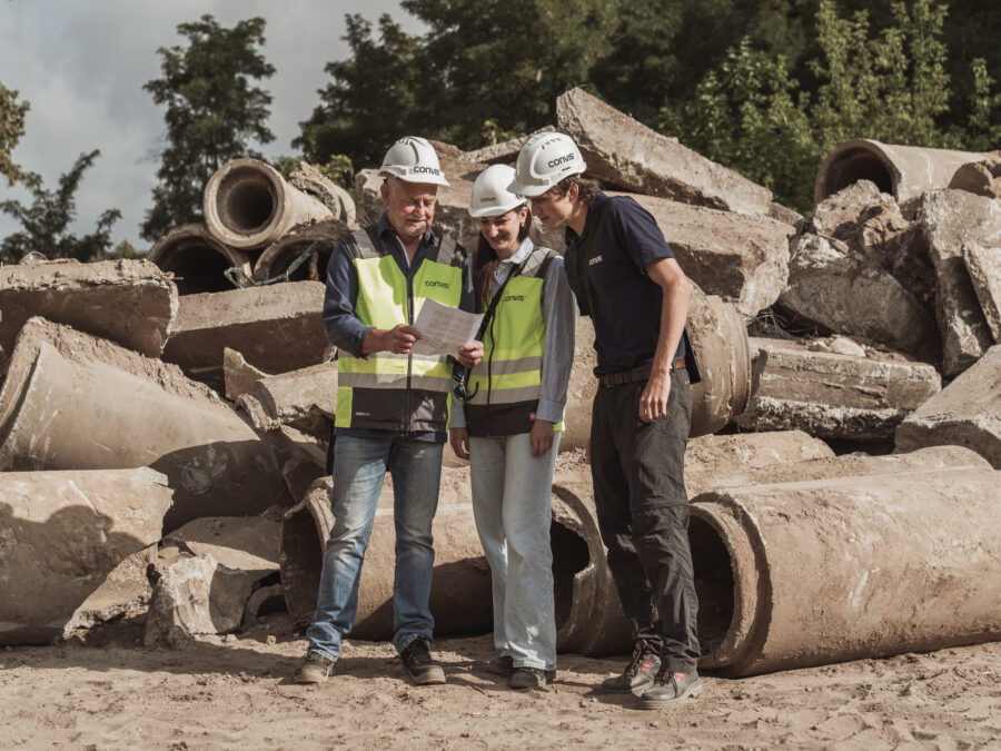 Bauarbeiter besprechen Pläne vor einer Baustelle mit Betonrohren im Hintergrund.