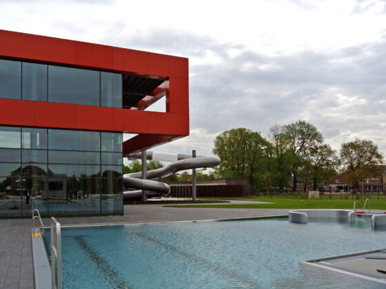 Moderne Schwimmbadanlage mit Rutsche, Glasfassade und großzügiger Grünfläche.