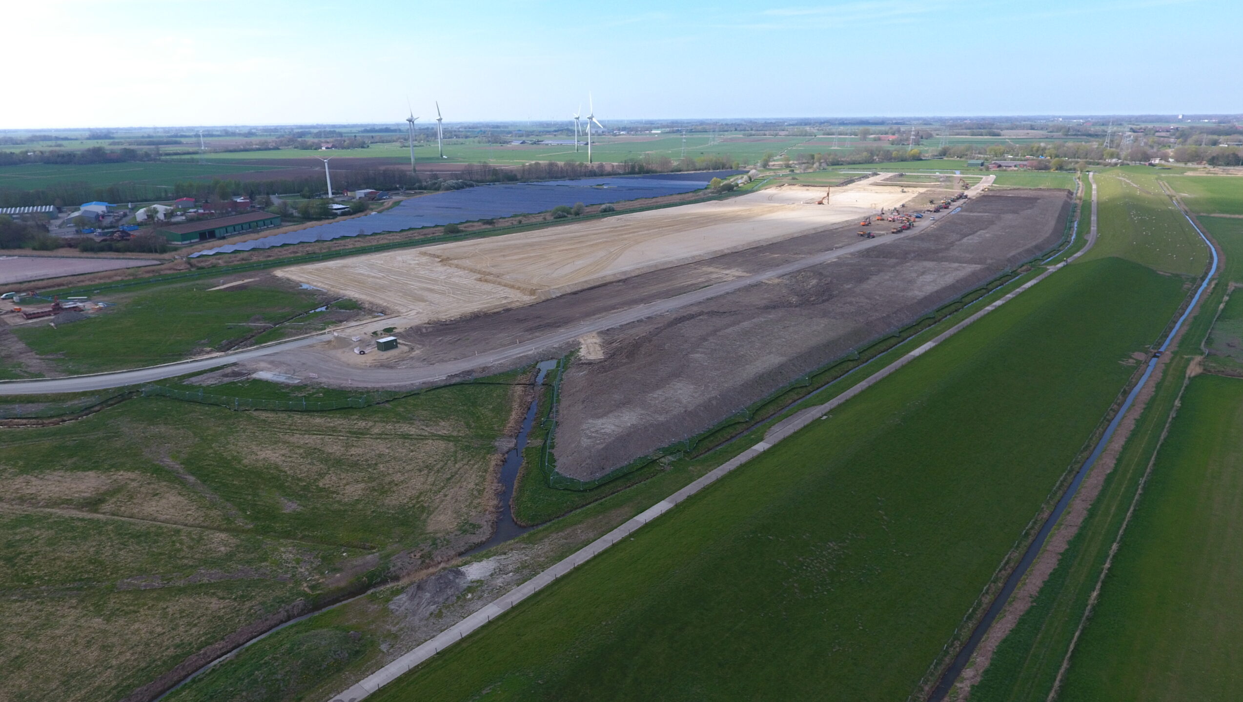 Bauarbeiten auf einem großen Grundstück mit Windkraftanlagen im Hintergrund.