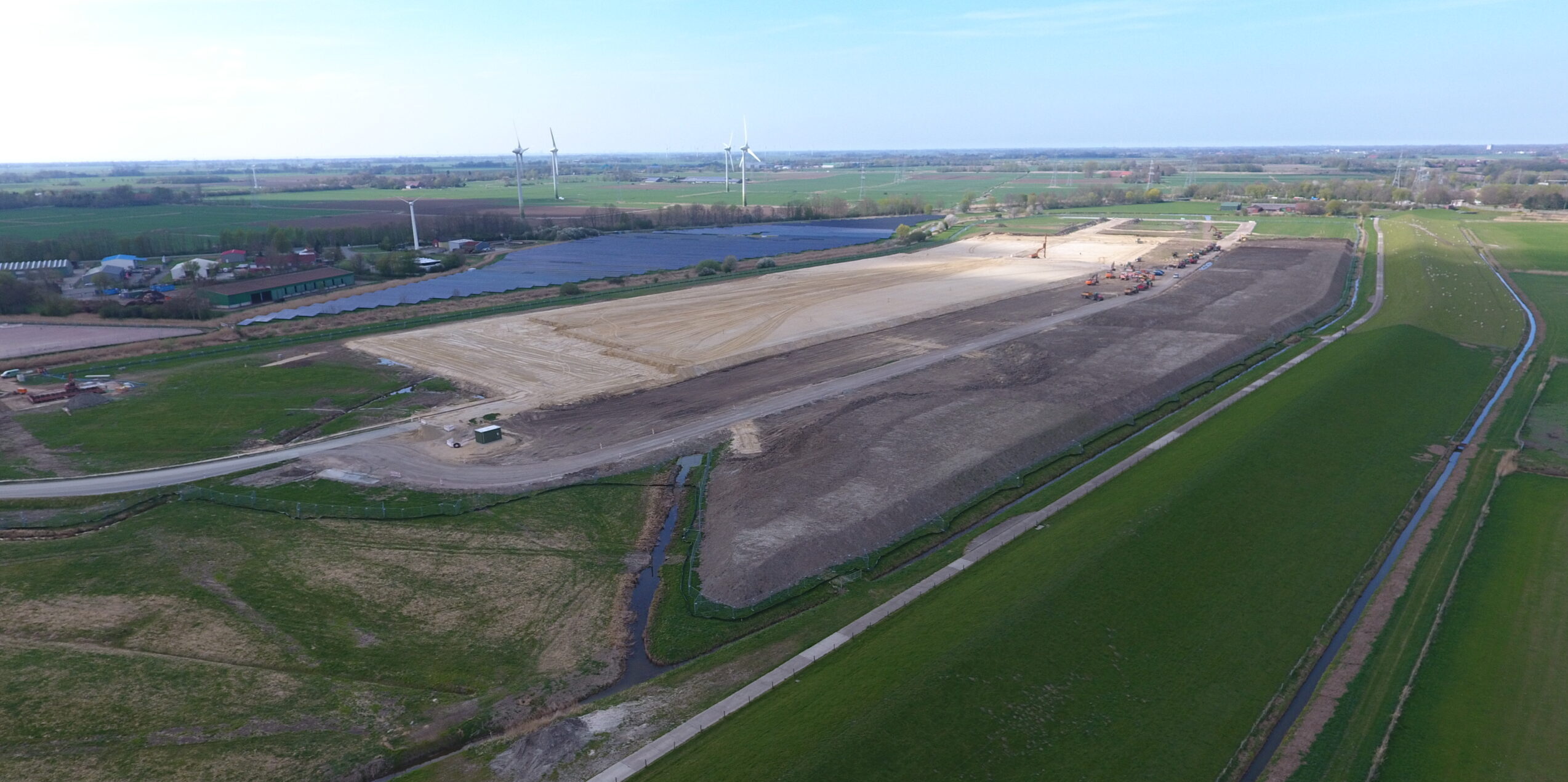 Bauarbeiten auf einem großen Grundstück mit Windkraftanlagen im Hintergrund.
