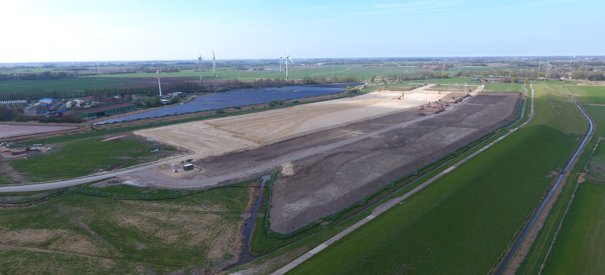 Bauarbeiten auf einem großen Grundstück mit Windkraftanlagen im Hintergrund.