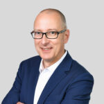 Professioneller Mann in Anzug mit Brille, lächelnd vor grauem Hintergrund.