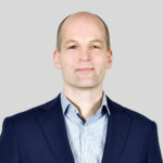 Professioneller Mann in Anzug vor neutralem Hintergrund, lächelnd und selbstbewusst.
