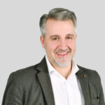 Professioneller Mann mit grauen Haaren lächelt in Anzug vor grauem Hintergrund.