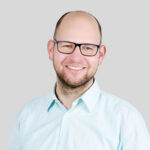 Lächelnder Mann mit Brille und hellblauer Hemd vor grauem Hintergrund.
