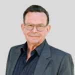 Geschäftsmann im Anzug mit Brille, lächelt vor grauem Hintergrund.