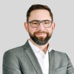 Professioneller Mann in Anzug mit Brille, lächelt vor grauem Hintergrund.