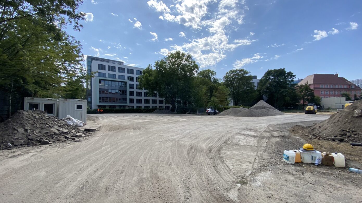 Bauplatz mit Erde und Bauschutt, umgeben von Bäumen und modernen Gebäuden.
