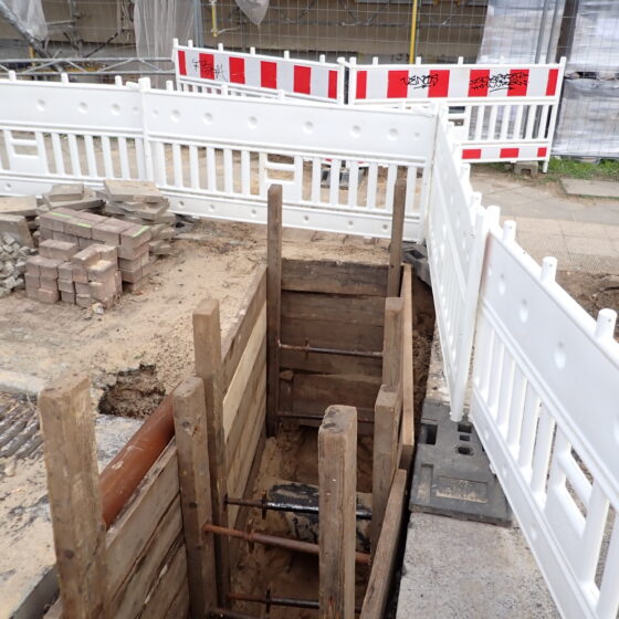 Baustelle mit Grube und Holzverkleidung, umgeben von Absperrungen und Baustellenmaterial.