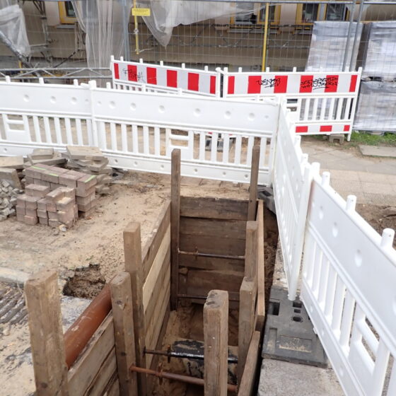 Baustelle mit Grube und Holzverkleidung, umgeben von Absperrungen und Baustellenmaterial.