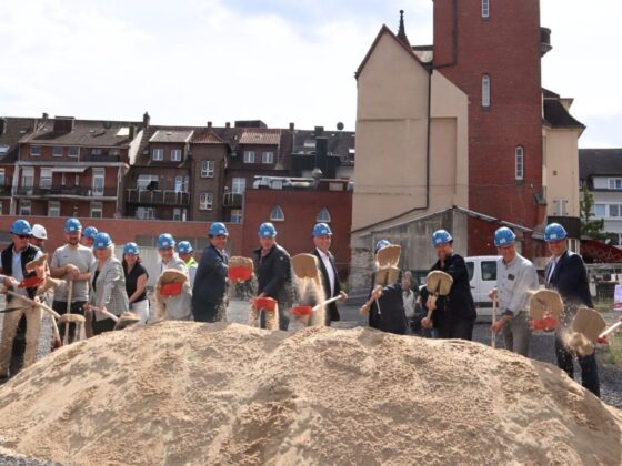 Feierliche Grundsteinlegung mit Bauhelmen und Schaufeln auf einer Baustelle.