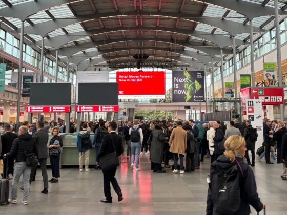 Menschenmenge auf einer Messe mit digitalen Anzeigen und moderner Architektur im Hintergrund.