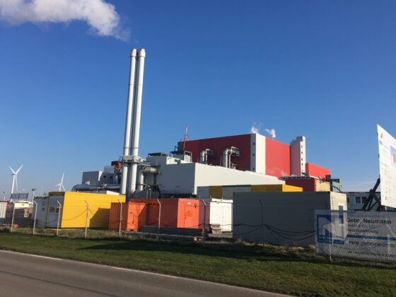 Industrieanlage mit Schornsteinen, Windrädern und bunten Containern vor klarem Himmel.