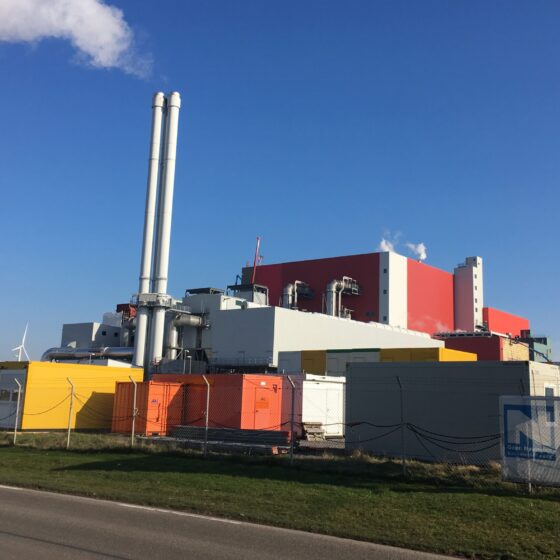 Industrieanlage mit Schornsteinen, Windrädern und bunten Containern vor klarem Himmel.